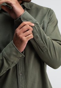 Olivengroene blouse met lange mouwen en een knoopsluiting aan de kraag, voorzien van een borstzak en verstelbare manchetten met knoopdetails. Zachte stofstructuur.