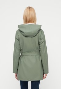 Γυναίκα με ξανθά μαλλιά που φοράει μια πράσινη βροχερή κάπα με κουκούλα και ζώνη, και σκούρα μπλε τζιν, εμφανιζόμενη από πίσω με φόντο ένα λευκό φόντο.