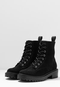 Botas de tobillo de ante negro con un acabado texturizado, cordones en la parte delantera, ojales de metal y una suela gruesa de goma negra para mayor tracción.