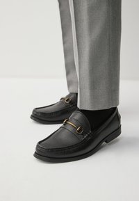 Mocassins en cuir noir avec finition texturée, détails en métal doré, et bout fermé. Associés à un pantalon gris sur mesure, présentés sur une surface blanche.