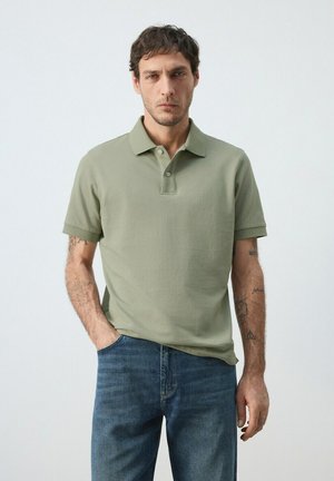 Homme aux cheveux courts et foncés portant un polo vert olive clair et un jean bleu, debout devant un fond clair uni.
