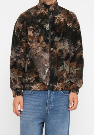 Homme portant une veste polaire à motif camouflage marron et noir avec une fermeture éclair à l'avant et un jean bleu sur un fond blanc uni.