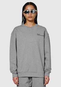 Grijze sweatshirt met lange mouwen, geribde manchetten en zoom. Heeft zwarte tekst "HALO 196819721981" op de borst. Draagt met een donkere zonnebril.