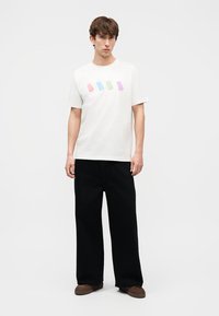 Νεαρός άνδρας που φοράει λευκό t-shirt με πολύχρωμα σχέδια παγωτού ξυλάκι, μαύρο παντελόνι φαρδιάς γραμμής και καφέ παπούτσια, σε απλό λευκό φόντο.