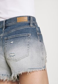 Džínové šortky s gradací od tmavě modré po světle modrou. Mají rozedrané lemy, vybledlé detaily a hnědou koženou nálepku s nápisem "Hollister."