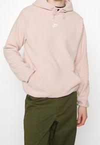 Felpa con cappuccio in pile rosa chiaro con cappuccio regolabile tramite coulisse e logo Nike bianco sul petto. Presenta una tasca a marsupio e polsini a costine.