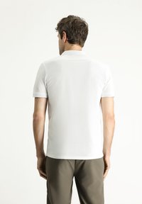 Polo shirt bianco a maniche corte con superficie testurizzata e colletto a coste, abbinato a pantaloni grigi chiari. Vista posteriore che mostra i dettagli del design.