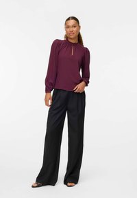 Blusa bordeaux a maniche lunghe con scollo arricciato e dettaglio a occhiello, abbinata a pantaloni neri a gamba larga e sandali neri piatti.
