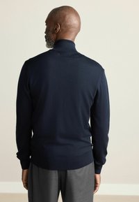 Marineblå, langærmet sweater med høj krave og ribbede manchetter. Teksturen virker glat og vises bagfra mod en lys baggrund.