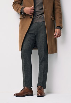 Homme portant un pardessus marron, un pull gris, un pantalon gris à motifs et des chaussures habillées en cuir marron, debout sur fond blanc.