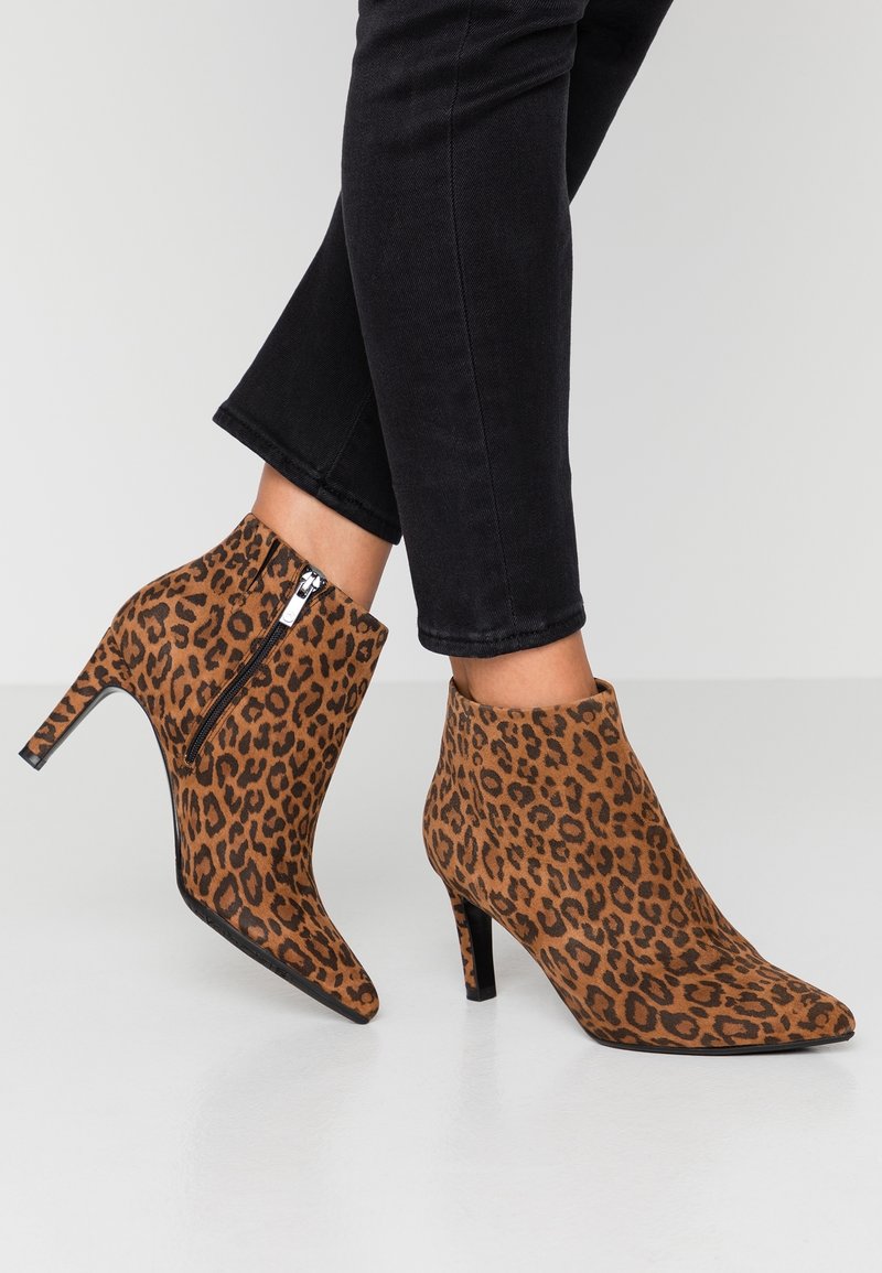 Bottes à talons aiguilles brun léopard montant à la cheville avec des fermetures éclair, portées avec un pantalon noir ajusté sur un sol blanc.
