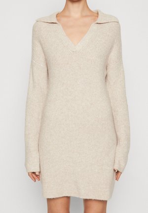 Beige gebreide jurk met lange mouwen, brede kraag en V-hals, gedragen door een persoon tegen een effen achtergrond.