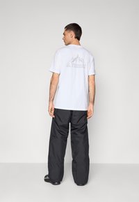 Camisa blanca con gráfico de montaña y logo "TERREX" en la parte posterior, combinada con pantalones negros de corte holgado y zapatos negros.