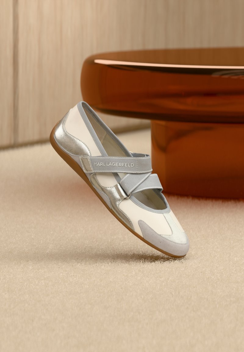 Λευκές και ασημένιε ς μπαλαρίνες με σταυρωτά λουράκια, με απαλό μείγμα υλικών και διακριτικό γυαλιστερό φινίρισμα, τοποθετημένες πάνω σε υφασμάτινη επιφάνεια.