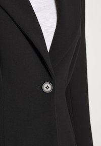 Gros plan sur un revers de blazer noir avec un seul bouton gris fermé sur un fond de chemise blanche.