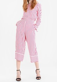 Vrouw in een rood-wit verticaal gestreept langadarig shirt en bijpassende cropped broek, met zwarte hoge hakken.