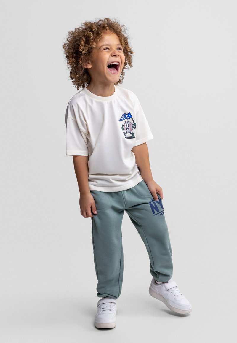 Enfant aux cheveux bouclés riant, portant un t-shirt blanc avec un dessin animé et un pantalon de survêtement gris avec des lettres bleues, se tenant sur un fond uni.