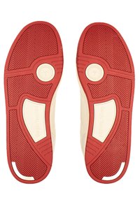 Podošvy topánok s červeným vzorovaným gripom a krémovo sfarbeným základom. Obsahujú kruhové logo a textúrované bočné akcenty.