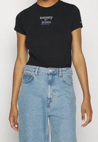 Svart bomullströja med "tommy jeans"-logotyp i vitt och blått, kombinerad med ljusblå denimjeans med hög midja och klassiska fickor.