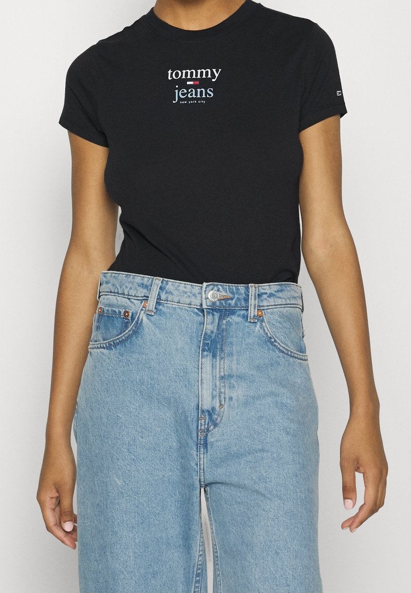 Svart bomullströja med "tommy jeans"-logotyp i vitt och blått, kombinerad med ljusblå denimjeans med hög midja och klassiska fickor.