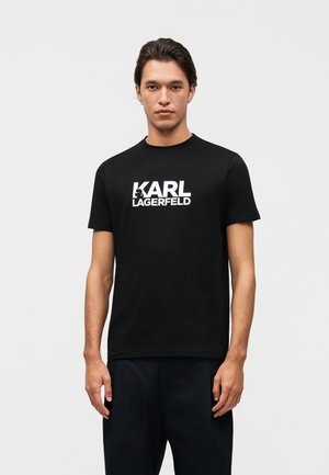 Mladić s tamnom kosom nosi crnu majicu kratkih rukava s bijelim natpisom "KARL LAGERFELD" u sredini na jednobojnoj pozadini.