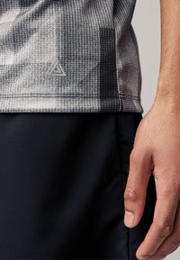 Graues Sportshirt mit Muster und strukturierter Oberfläche, Logodetail nahe dem Saum, kombiniert mit schwarzen Shorts. Hand ruht sanft auf den Shorts.