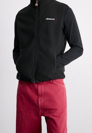 Person trägt eine schwarze Fleeceweste mit Reißverschluss über einem langärmligen schwarzen Shirt und roten Hosen, die Hände sind in den Taschen der Weste.