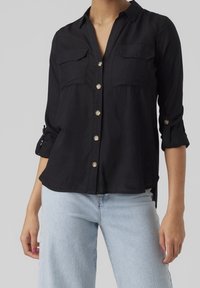 Camicia nera con bottoni, colletto, due tasche frontali e maniche arrotolate. Realizzata in tessuto leggero con una texture liscia.