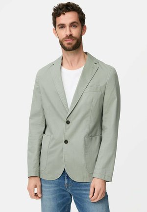 Mann trägt hellgrünen Freizeitblazer über weißem Hemd und blauer Jeans, steht vor einfarbigem Hintergrund.