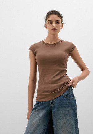 Donna che indossa una maglietta marrone aderente a coste con maniche corte e jeans blu larghi, in piedi davanti a uno sfondo grigio chiaro semplice.