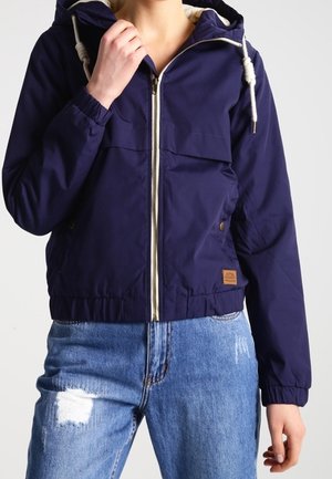 Vrouw die een marineblauwe capuchonvest met ritssluiting draagt met witte trekkoorden en lichtblauwe gescheurde spijkerbroek, staand tegen een effen achtergrond.