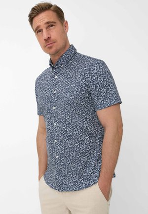 Mand iført en kortærmet navyblå skjorte med hvide bladprint og beige bukser, stående med hænderne i lommerne.