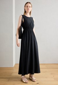 Abito maxi senza maniche di colore nero con vita elasticizzata, abbinato a sandali con tacco a blocco dorati e cinturini. Lo sfondo presenta un pavimento in legno chiaro.