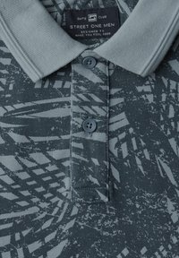 Polo en gris oscuro con un patrón geométrico en gris más claro. Presenta un cuello suave, tapeta de tres botones y una tela texturizada.