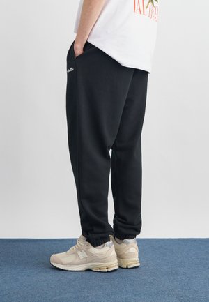 Osoba nosząca czarne spodnie dresowe z elastycznymi mankietami oraz beżowe buty New Balance, stojąca na niebieskim dywanie.