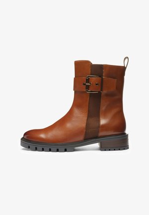 Brown leather ankle boot with black rubber lug sole, side elastic panel, decorative strap with buckle, and pull tab at the back.