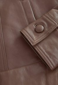 Vêtement en cuir marron avec des coutures détaillées et un gros plan d'un poignet sécurisé par un bouton rond.