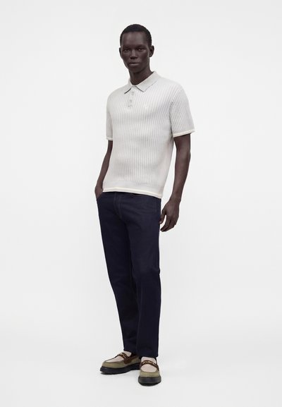 Homme debout portant un polo blanc texturé à manches courtes, un pantalon foncé et des mocassins bicolores sur un fond blanc uni.