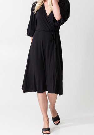 Kobieta w czarnej midi sukience kopertowej z bufkami na rękawach oraz czarnych, otwartych sandałach na płaskiej podeszwie, stojąca na tle jednolitego tła.