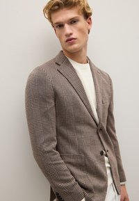 Braun karierter Blazer aus strukturiertem Stoff mit schlankem Schnitt, ausgestattet mit einem einzigen Knopf und Revers, getragen über einem cremefarbenen Pullover.