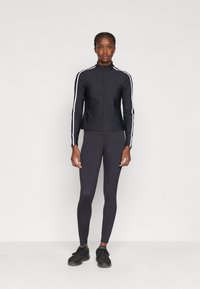 Svart atletisk zip-up jakke med hvite sidestriber, kombinert med mørke leggings og svarte sko. Glatt tekstur, tettsittende design.