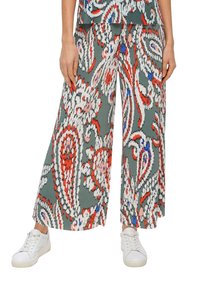 Pantalon à jambes larges avec des motifs floraux abstraits verts, blancs, rouges, bleus et roses, porté avec des baskets blanches sur une personne debout.