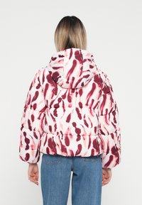 Geacă puf în alb cu un model abstract roșu și roz, având glugă, textură matlasată și un design scurt, asortată cu blugi albaștri.