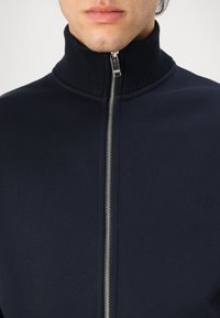 Marineblå zip-jakke med høy ribbestrikket krage og sølvglidelås. Myk tekstur, tettsittende design, med logo-detalj på brystet.