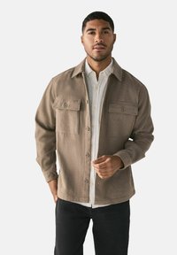 Ruskea villasekoitepaitatakki, jossa on kaksi etutaskua, nappikiinnitys ja kaulus, päällä valkoinen nappikauluspaita ja tummat farkut.