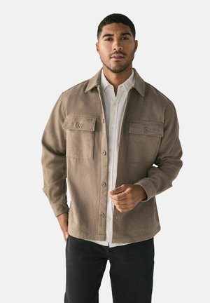 Braune Wollmischung-Hemddjacke mit zwei vorderen Taschen, Knopfschluss und Kragen, getragen über einem weißen Hemd mit Knöpfen und dunkler Jeans.