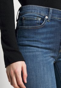 Jeans de mezclilla azul con un ligero degradado, que cuentan con cinco bolsillos, costuras amarillas y una cintura alta. La textura es suave y ajustada.