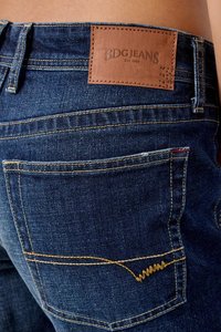 Zbliżenie na tylną kieszeń ciemnoniebieskich dżinsów z denimu z brązową skórzaną łatką z logo marki BDG JEANS EST 1998.