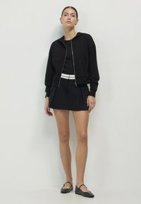 Un hoodie noir court avec une fermeture éclair argentée, associé à une jupe plissée noire et des chaussures plates. L'ensemble est rehaussé d'une ceinture blanche.