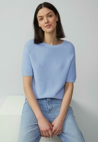 Tunique en tricot bleu clair avec des manches raglan courtes, un col rond et une finition texturée, associée à un jean en denim bleu clair.